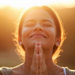 Eine Frau lächelt mit geschlossenen Augen, hält die Hände in Gebetshaltung und genießt den Sonnenuntergang im Hintergrund.