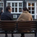 Zwei Personen sitzen auf einer Holzbank und unterhalten sich, mit einer Häuserfassade im Hintergrund