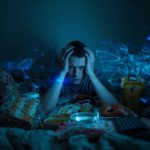 Ein junger Mann sitzt nachts in einem dunklen Raum zwischen vielen Snacks und Fast Food, hält seinen Kopf verzweifelt in den Händen, während auf einem Bildschirm eine Uhr kurz vor Mitternacht zeigt.