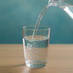 Wasser wird aus einer Glaskaraffe in ein halb gefülltes Trinkglas auf einem Holztisch gegossen.