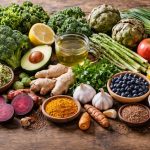 Auswahl frischer, pflanzlicher Lebensmittel wie Brokkoli, Spargel, Ingwer, Kurkuma, Beeren, Knoblauch, Artischocken, Zitronen, Nüsse und grüner Tee auf einem Holztisch, angerichtet in natürlichem Licht.