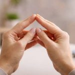 Zwei Hände formen mit den Fingerspitzen ein Dreieck und sind mittig im Bild vor einem weich unscharfen, hellen Arbeitsumfeld zu sehen.