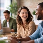Zwei Kolleg:innen sitzen an einem Tisch im Büro und lachen miteinander, während im Hintergrund weitere Teammitglieder angespannt arbeiten.