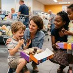 Zwei Frauen und zwei Kinder probieren lachend Schokoküsse in einem Supermarkt, neben ihnen steht ein Einkaufswagen mit Lebensmitteln.