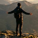 Mann mit Rucksack steht auf einem Berggipfel mit ausgebreiteten Armen und blickt über eine weite, bergige Landschaft im warmen Licht.