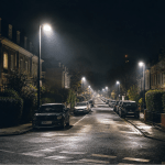 Eine nächtliche Wohnstraße mit modernen LED-Straßenlaternen, deren kaltes, helles Licht stark in die Fenster eines Hauses fällt, während eine ältere Frau im Innenraum sichtbar unter der Helligkeit leidet.