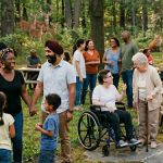 Eine vielfältige Gruppe von Menschen unterschiedlichen Alters und Hintergrunds trifft sich in einem Waldpark zu einem gemeinsamen Picknick. Kinder spielen im Vordergrund, eine Person sitzt im Rollstuhl, und eine ältere Frau steht mit Gehstock daneben. Im Hintergrund sind Wildtiere wie Rehe, Füchse und Truthähne zwischen den Bäumen zu sehen.