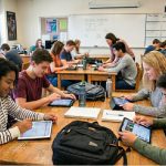 Schüler sitzen in einem hellen Klassenraum an Holztischen und arbeiten gemeinsam mit Tablets und Laptops.