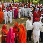 Große Menschenmenge bei einer traditionellen Zusammenkunft im Freien. Viele Personen tragen weiße Gewänder und Kopfbedeckungen, während zahlreiche Frauen farbenfrohe Saris in Rot-, Orange-, Pink- und Gelbtönen tragen. Die Menschen stehen in Gruppen zwischen Bäumen und unterhalten sich.