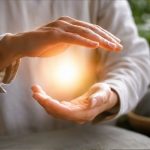 Eine Person hält die Hände vor dem Körper in einer meditativen Haltung. Zwischen den Handflächen scheint ein sanftes, goldenes Licht zu schweben. Im Hintergrund sind weiche, harmonische Lichtstrukturen und subtile geometrische Muster zu erkennen, die Ruhe, Energiefluss und Achtsamkeit symbolisieren. Die Szene wirkt spirituell, friedlich und inspirierend.