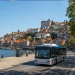 Moderner Linienbus fährt auf einer breiten Straße entlang des Douro-Ufers in Porto. Im Hintergrund sind die historische Altstadt, zahlreiche Fußgänger und eine grüne Uferpromenade bei sonnigem Wetter zu sehen.