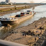 Ein Frachtschiff fährt bei niedrigem Wasserstand durch ein begradigtes Flussbett mit befestigten Ufern, während am freiliegenden Kiesufer Menschen entlanggehen.