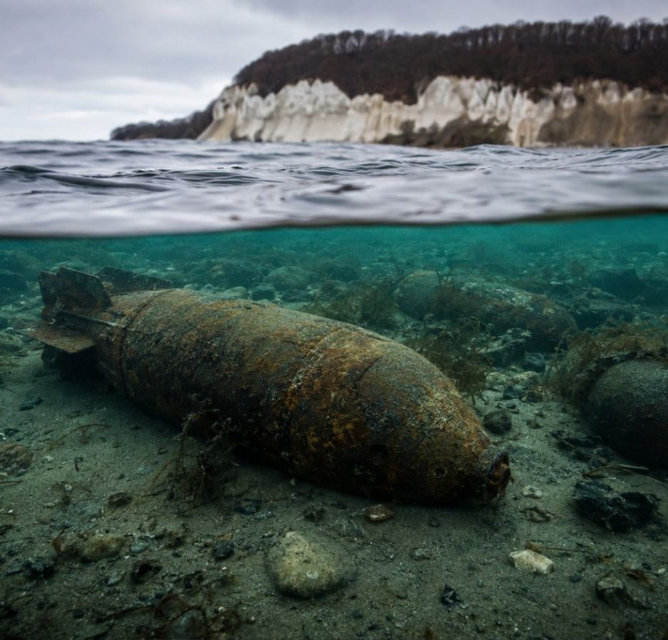 Unterwasseraufnahme mit stark verrosteter Weltkriegsmunition auf dem Meeresgrund der Ostsee; im Hintergrund sind die Küste und die Wasseroberfläche zu sehen.