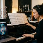 Eine Frau sitzt mit Kopfhörern an einem Klavier in einem Musikstudio. Auf einem Notenständer liegen Notenblätter mit DNA-Sequenzen, während auf einem Laptop-Bildschirm Schwingungsfrequenzen und eine leuchtende DNA-Doppelhelix angezeigt werden. Im Hintergrund stehen ein DNA-Modell und Bücherregale.