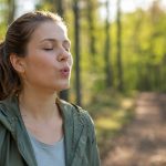 Junge Frau pfeift entspannt mit geschlossenen Augen in einem sonnigen Wald.