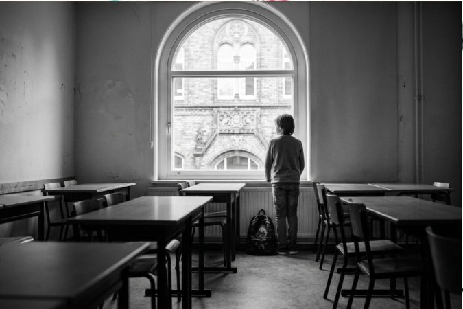 Ein Kind steht von hinten gesehen alleine in einem leeren Klassenzimmer an einem großen Rundbogenfenster und blickt nach draußen auf ein historisches Gebäude.